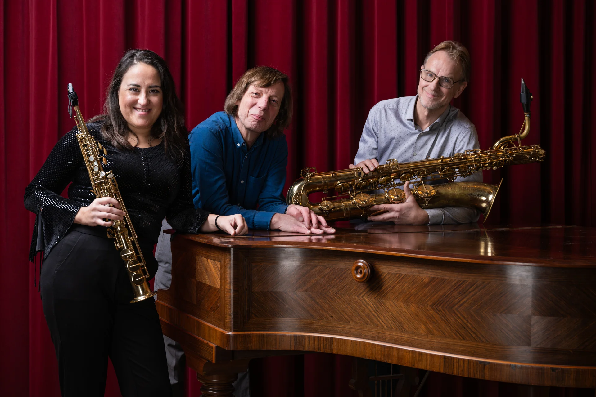 The three musicians of Trio Abril with two saxophones beside a grand piano, in front of a red theatre curtain.