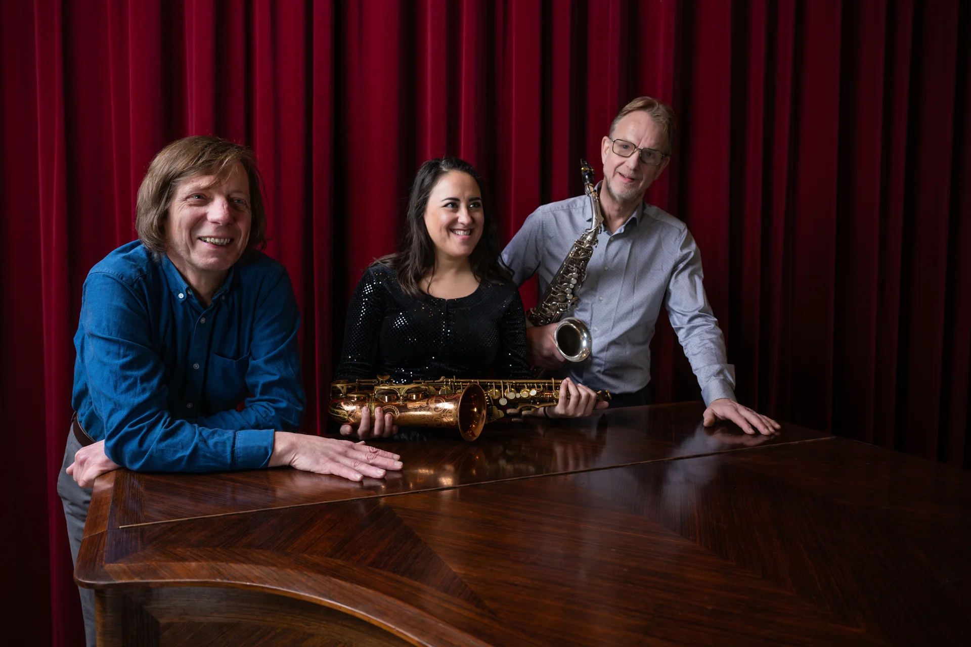The three musicians of Trio Abril with two saxophones beside a grand piano, in front of a red theatre curtain.